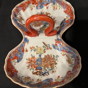 May include: A white porcelain serving dish shaped like a seashell with a red handle. The dish is decorated with a floral pattern in shades of red, blue, gold, and green.