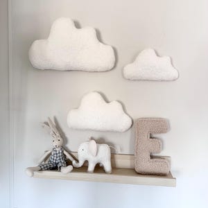 May include: A nursery wall display with three fluffy white cloud cushions, a wooden shelf holding a stuffed bunny and elephant, and a tan letter "E". The clouds are fixed to a white wall.