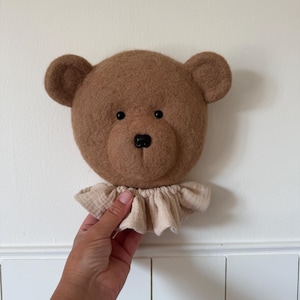 May include: A brown felted bear head wall decoration featuring two rounded ears, small black bead eyes, and a black nose. The bear has a ruffled, light beige fabric collar attached around the neck area, creating a soft and textured appearance.