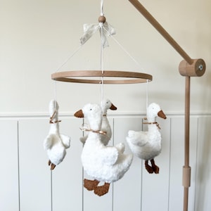 Puede incluir: Móvil para bebé de madera con cuatro gansos blancos de peluche, picos y patas marrones. Los gansos están suspendidos de un anillo y un brazo de madera. Un lazo decorativo está atado en la parte superior del móvil.