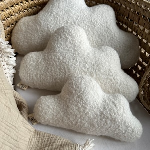 May include: Three white cloud-shaped decorative pillows with a textured, fluffy surface. The pillows are arranged in a wicker basket with a beige blanket. Each pillow has a small tag.
