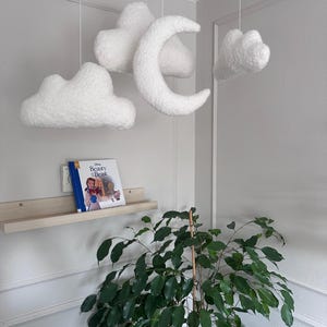 Puede incluir: Móvil de nubes y luna creciente blancas y esponjosas colgando del techo. La decoración de la guardería incluye una pequeña estantería de madera con el libro "Beauty and the Beast" y una planta verde.