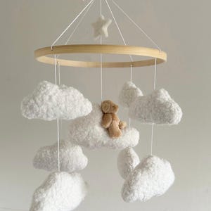 May include: A baby mobile featuring a wooden ring, a star, and fluffy white clouds. A small teddy bear sits atop one of the clouds. The mobile is suspended by white strings, creating a whimsical nursery decoration.