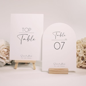 Può includere: Due segnaposto bianchi con testo nero. Uno recita "TOP Table" ed è su un piccolo cavalletto di legno. L'altro, ad arco, recita "Table NO 07" ed è su una base di legno. Entrambi i segnaposto riportano i nomi "Simon & Alexis" e la data "21.09.2024".