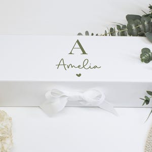 May include: A rectangular white gift box featuring the letter A and the name Amelia printed in olive green, with a small olive green heart icon below the name and a white ribbon tied in a bow at the center of the front panel.