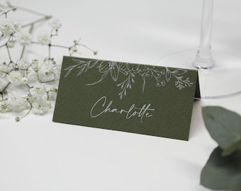 May include: An olive green place card, folded and bearing the name "Charlotte" in white script. The top edge is decorated with white floral line art. The card sits on a white surface, with white flowers and a wine glass visible.
