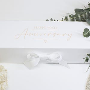 Könnte beinhalten: Eine rechteckige weiße Geschenkbox mit dem goldenen Schriftzug HAPPY 60TH Anniversary und einem kleinen Herzsymbol. Die Box ist mit einer weißen Schleife in der Mitte verschlossen. Das Design ist schlicht und einfach mit einer glatten, matten Oberfläche.