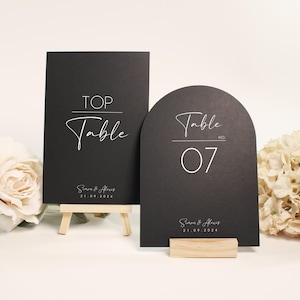 Può includere: Due segnaposto neri con calligrafia bianca. Uno è rettangolare e recita "TOP Table". L'altro è arcuato e dice "Table No. 07". Entrambi includono nomi e una data. I segnaposto sono su supporti in legno, con fiori sullo sfondo.