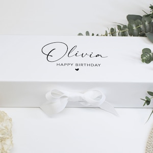 Può includere: Una scatola regalo rettangolare bianca con il nome Olivia in un carattere corsivo nero, seguito dal testo HAPPY BIRTHDAY e da una piccola icona a forma di cuore. La scatola è chiusa con un nastro bianco annodato al centro del lato frontale.