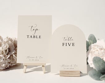 May include: Two ivory wedding table signs. One rectangular sign on a small easel reads "Top Table". The other, arched sign reads "Table FIVE". Both signs include the names "Harrison & Eva" and the date "21.09.2024".