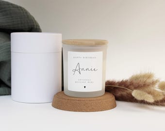 Puede incluir: Una vela de vidrio esmerilado con tapa de madera, etiquetada como "Happy Birthday Annie". La vela está sobre un posavasos de corcho, junto a una caja cilíndrica blanca. Flores secas y una tela verde en el fondo.