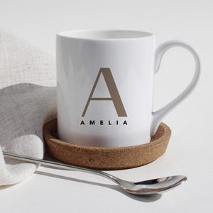 Könnte beinhalten: Weiße Keramiktasse mit braunem Buchstaben "A" und dem Namen "AMELIA" aufgedruckt. Die Tasse steht auf einem Korkuntersetzer, mit einem Löffel und einem Leinentuch im Hintergrund. Schlichtes, elegantes Design.