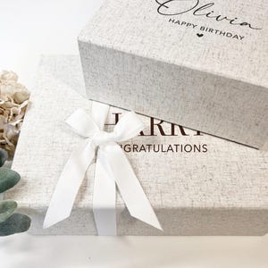 Puede incluir: Dos cajas de regalo rectangulares con una textura gris claro similar al lino. La caja superior presenta el texto Olivia HAPPY BIRTHDAY con un corazón. La caja inferior está atada con una cinta de satén blanca y presenta el texto CONGRATULATIONS.