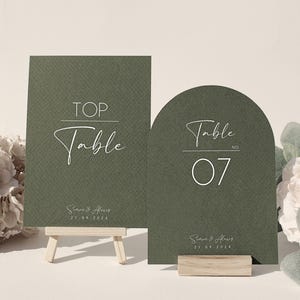 Può includere: Due segnaposto verde oliva con testo bianco. Uno è rettangolare e recita "TOP Table". L'altro è ad arco e recita "Table No. 07". Entrambi i segnaposto includono nomi e una data. Sono esposti su piccoli supporti in legno.