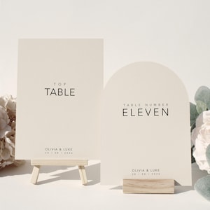 Può includere: Due segnaposto per tavoli da matrimonio color crema. Un segnaposto rettangolare su un piccolo cavalletto di legno con la scritta "TOP TABLE". L'altro, un segnaposto ad arco con la scritta "TABLE NUMBER ELEVEN". Entrambi i segnaposto hanno nomi e una data.