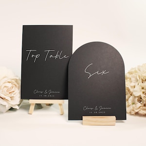 Può includere: Due segnaposto neri per matrimoni. Un segnaposto rettangolare con la scritta "Top Table" è appoggiato su un piccolo cavalletto di legno. L'altro, a forma di arco, recita "Six". Entrambi i segnaposto riportano i nomi "Chris & Joanna" e la data "11.10.2025".