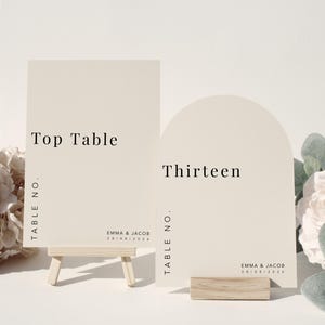 Può includere: Due segnaposto con numeri da tavolo bianchi. Un segnaposto rettangolare recita "Top Table" ed è sostenuto da un piccolo cavalletto di legno. L'altro segnaposto è ad arco e recita "Thirteen". Entrambi i segnaposto hanno il testo "Emma & Jacob" e una data.