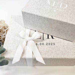 Puede incluir: Dos cajas rectangulares de color gris claro texturizado con lazos de cinta blanca. La caja superior presenta el texto blanco M | D y 14TH JUNE 2026 con un corazón. La caja inferior presenta el texto negro L | R y 6.08.2025. Aspecto de lino.