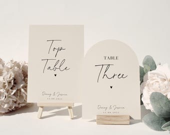 May include: Two cream-colored wedding table signs. One rectangular sign reads "Top Table" and the other, arched sign reads "Table Three". Both signs include names and a date. The signs are on wooden stands.