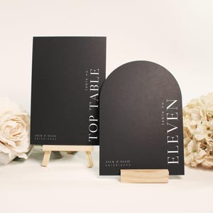 Puede incluir: Dos carteles negros con números de mesa con texto blanco. Uno es rectangular y dice "TOP TABLE", el otro es arqueado y dice "ELEVEN". Ambos tienen nombres y una fecha en la parte inferior. Se muestran con flores.