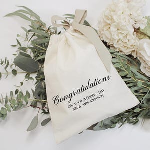 May include: A cream-colored fabric drawstring pouch featuring the black printed text Congratulations ON YOUR WEDDING DAY MR & MRS JOHNSON. The bag is tied at the top with a matching cream ribbon and is positioned against a backdrop of green eucalyptus leaves and white flowers.