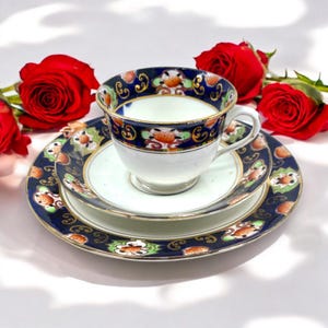 Può includere: Un servizio da tè vintage con tazza, piattino e piatto. Il set presenta un bordo blu scuro con finiture dorate e motivi floreali. L'interno della tazza e del piattino è bianco. Rose rosse sullo sfondo.
