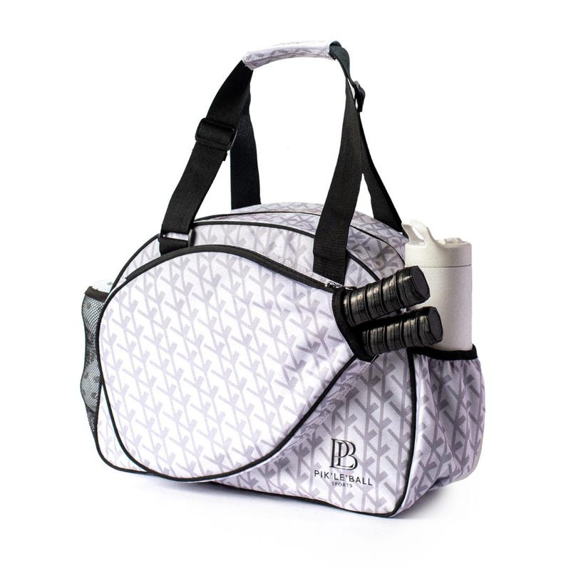 K&ouml;nnte beinhalten: Eine wei&szlig;e und graue Pickleball-Tasche mit sich wiederholendem Muster. Die Tasche hat schwarze Tr&auml;ger und Griffe, mit einer Seitentasche, die zwei Pickleball-Schl&auml;ger und eine Wasserflasche enth&auml;lt. Auf der Tasche steht der Text "PIK'LE BALL SPORTS".