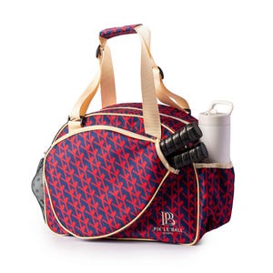 Könnte beinhalten: Pickleball-Tasche mit marineblauem und rotem Muster mit cremefarbenen Akzenten. Die Tasche hat einen cremefarbenen Griff und Schultergurt. Zwei Pickleball-Schläger und eine weiße Wasserflasche befinden sich in den Seitentaschen. Die Tasche trägt den Text "PIK'LE BALL SPORTS".