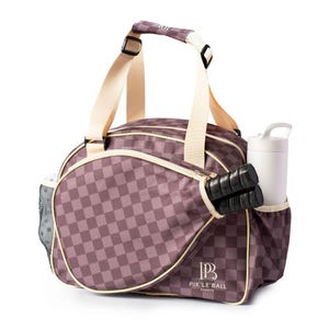 K&ouml;nnte beinhalten: Pickleball-Tasche mit einem karierten Muster in Lila- und Cremet&ouml;nen. Die Tasche hat ein Rei&szlig;verschluss-Hauptfach, eine Seitentasche f&uuml;r eine Wasserflasche und eine Tasche f&uuml;r Pickleball-Schl&auml;ger. Creme-farbener Griff und Schultergurt. Der Text "PIK'LE BALL SPORTS" ist sichtbar.