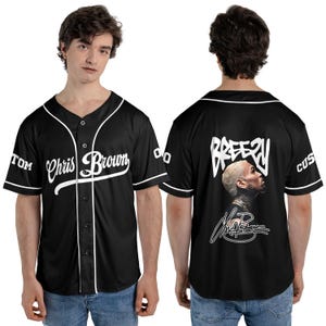 May include: Black baseball jersey with white trim and the name "Chris Brown" in script across the front. The back features the word "Breezy" above a portrait and signature. Sleeves have "TOM" and "CUS" text.