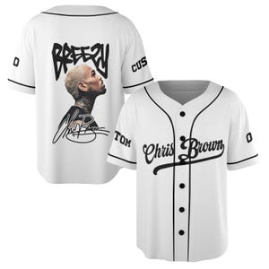 May include: White baseball jersey with black trim and buttons. The front features the text "Chris Brown" in a script font. The back has the word "Breezy" above a portrait and signature. The sleeves have the word "Cus" and the number "0".