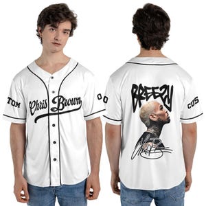 May include: White baseball jersey with black trim, featuring the text "Chris Brown" in script on the front. The back displays "Breezy" above a portrait and signature. Sleeves have "TOM" and "CUS" text.