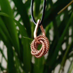 May include: A copper-coloured pendant necklace with a woven design. The pendant hangs from a black cord against a backdrop of green foliage. The pendant's woven design is a prominent feature.