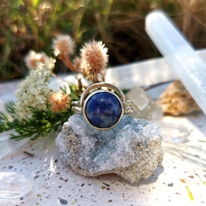 Peut inclure: Une bague en argent ornée d'une grosse pierre ronde bleu foncé. La bague est présentée sur une formation de cristal bleu clair, avec un arrière-plan flou de verdure et d'autres cristaux. Le design de la bague met en valeur la pierre.