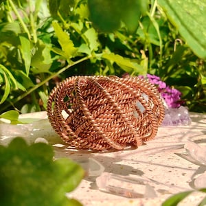 May include: A copper-coloured, woven bracelet rests on a light-coloured surface, surrounded by green foliage and small purple flowers. The bracelet is made of tightly wound copper wire, creating a textured, open design.