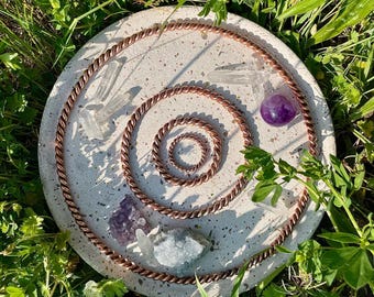 May include: A white circular tray with a copper wire design, featuring a variety of crystals, including clear quartz, amethyst, and a purple crystal. The tray is surrounded by green grass.