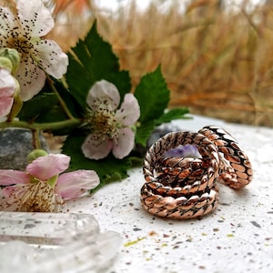 May include: Three copper rings stacked on top of each other. The rings are braided and have a shiny finish. The rings are on a white surface with a few small brown spots.