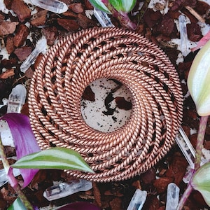 May include: A coiled copper-coloured spiral, possibly a decorative item, is the central feature. It's set amongst clear quartz crystals, purple and green leaves, and brown wood chips, creating a natural, earthy aesthetic.