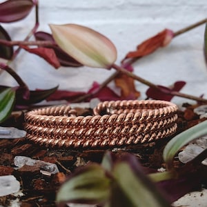 May include: Two copper-coloured bracelets with a twisted rope design. The bracelets are displayed on a bed of wood chips, with clear crystals and leafy plants in the background. The bracelets are a warm, metallic tone.