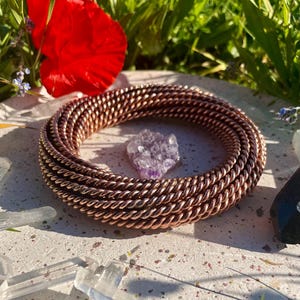 Könnte beinhalten: Ein kupferfarbenes, gewickeltes Drahtarmband umschließt einen kleinen, lila Amethystkristall. Das Armband liegt auf einer gesprenkelten Oberfläche, mit einer roten Blume und grünem Laub im Hintergrund. Klare Quarzkristalle sind verstreut.
