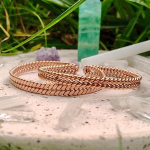Peut inclure: Bracelets de couleur cuivre, de style tressé, reposent sur une surface texturée de couleur claire, entourés de cristaux et de verdure. Un cristal vert et d'autres cristaux sont en arrière-plan. Les bracelets ont un design tissé.