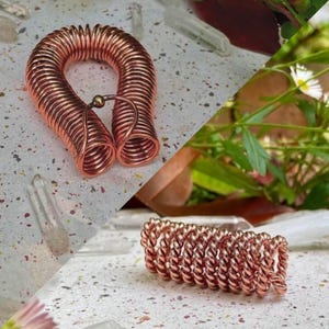 Könnte beinhalten: Zwei Kupferdrahtspulen, eine U-förmige und die andere eine enge Spirale. Die U-förmige Spule hat oben eine kleine goldfarbene Perle. Die Spulen werden auf einer gesprenkelten weißen Oberfläche mit einem Hintergrund aus Pflanzen und Kristallen gezeigt.