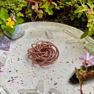 May include: A copper-coloured, twisted wire spiral sits on a speckled, light grey surface, surrounded by clear quartz crystals, small flowers, and greenery. The spiral is the focal point, with a detailed, woven appearance. The overall aesthetic is natural and earthy.