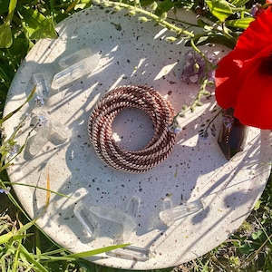 May include: A round, speckled concrete tray displays a coiled copper wire, clear quartz crystals, and small purple amethyst clusters. A bright red poppy and greenery surround the arrangement, giving a natural, earthy feel.