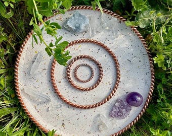 May include: A white circular tray with a copper wire design around the edge. The tray has a smaller copper wire circle in the center. There are several clear quartz crystals and amethyst crystals on the tray.