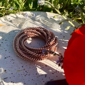 May include: A coiled copper bracelet with a twisted rope design. The bracelet is a warm, reddish-brown colour and sits on a speckled, light-coloured surface. The background includes green grass, small blue flowers, and a red flower.
