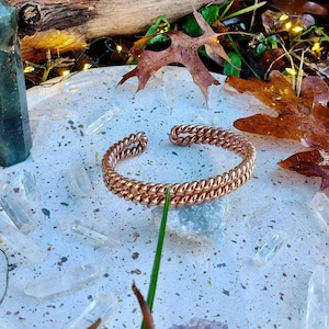 Pode incluir: Uma pulseira de punho cor de cobre com um design de corrente dupla. A pulseira é exibida em uma superfície salpicada de cor clara, cercada por cristais, folhas e um pedaço de madeira.