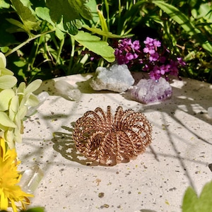 May include: A copper wire sphere sculpture rests on a textured surface, with crystals, purple flowers, and green foliage in the background. The sculpture is made of tightly wound copper wire.