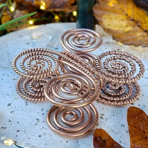 May include: A copper-coloured, spiral-shaped metal art piece with a green crystal point in the background. The artwork is set on a textured surface with fallen leaves and greenery, creating a natural aesthetic.