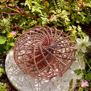 May include: A copper-coloured, woven wire sphere sits on a white, speckled, circular stone base. The sphere has a spiral design. The base has several clear crystal points. The background is filled with green foliage and small pink and white flowers.